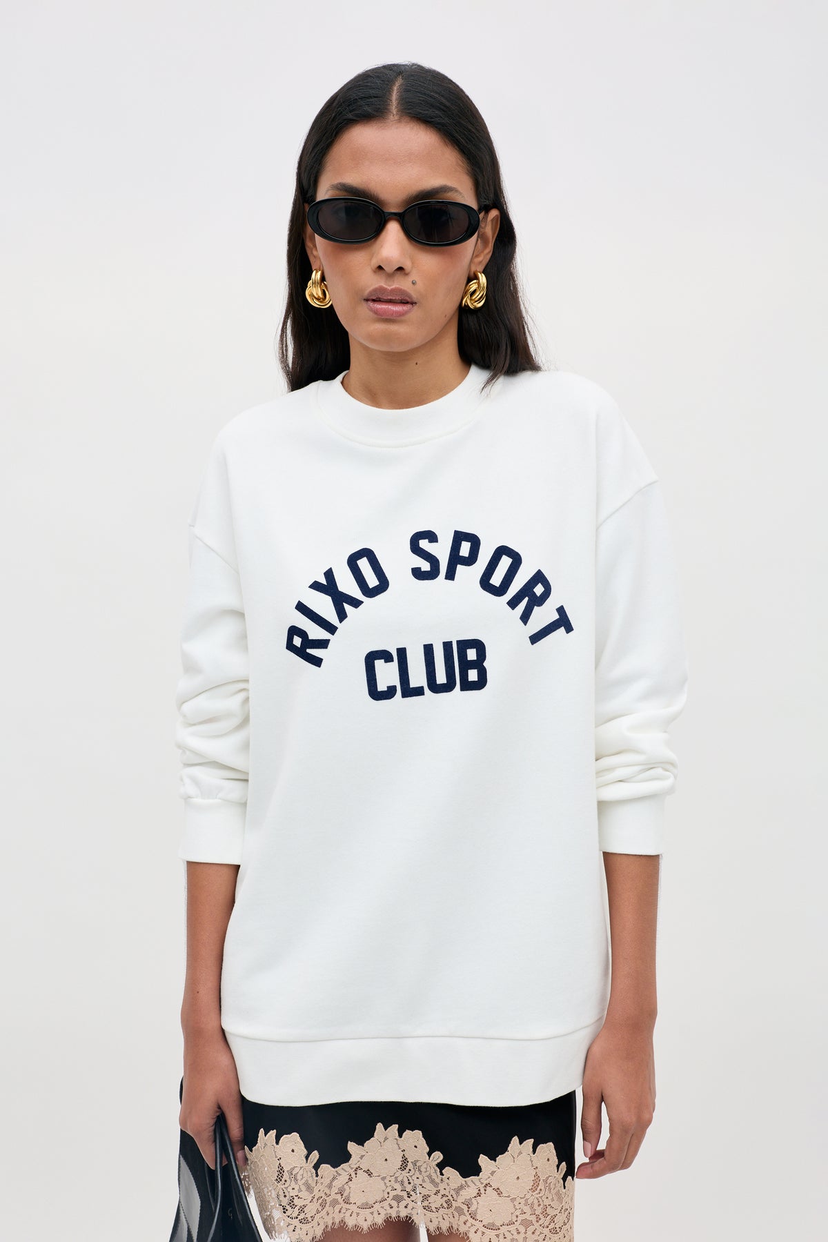 A woman wearing sunglasses and gold earrings models the Blaze - Rixo Sport Club White sweatshirt, paired with a lace-trimmed skirt and holding a bag.
