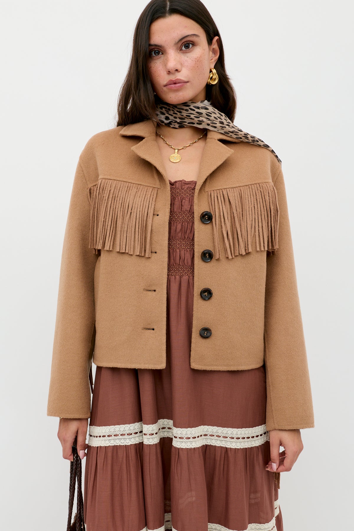 A woman wears the Ashleigh - Camel jacket with fringe over a mauve lace-accented dress, accessorized with a leopard print scarf and layered gold necklaces, standing against a plain white background.