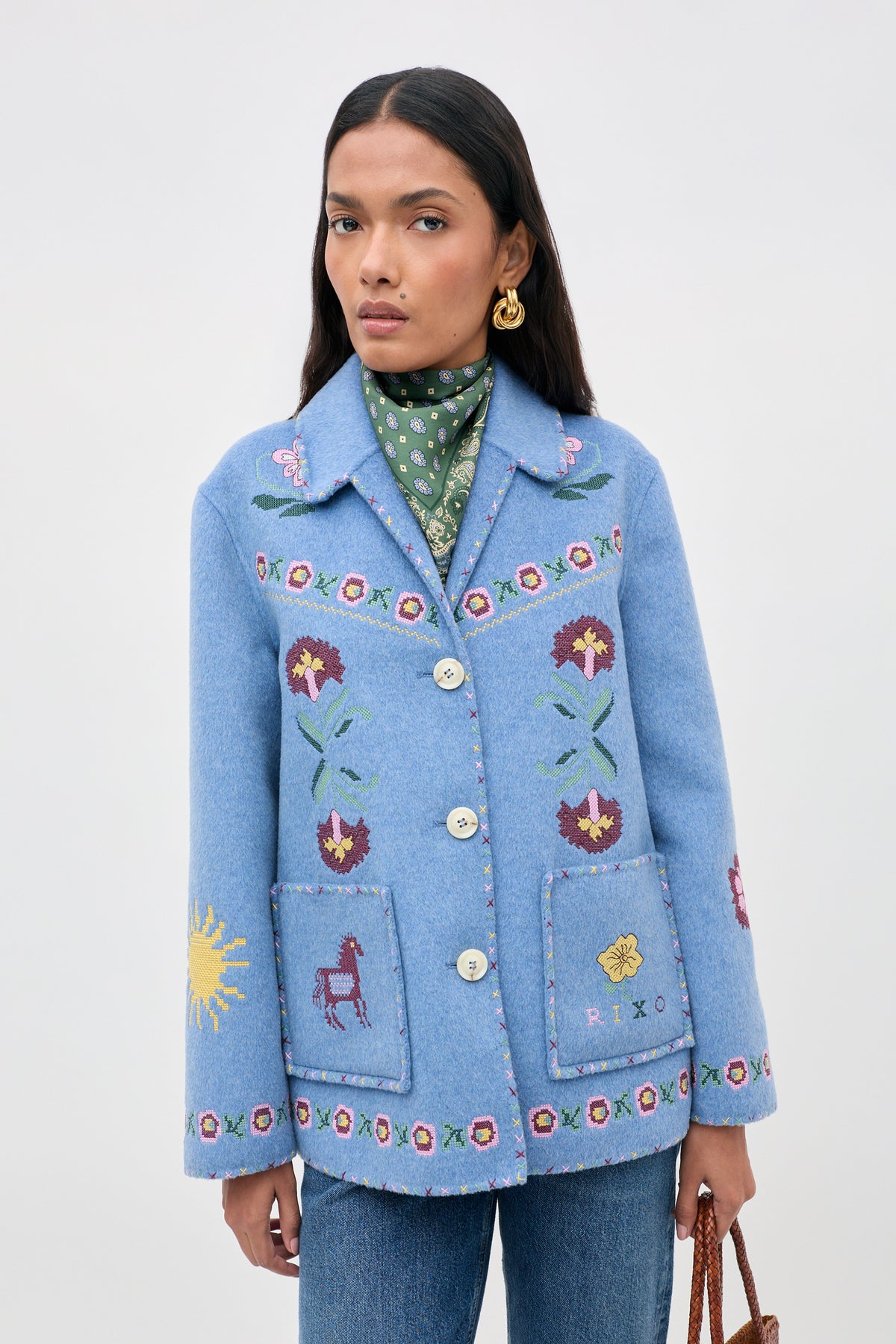 Wearing the Tashia - Juliette Embroidery Blue jacket adorned with floral and animal patterns, along with gold earrings and a green patterned scarf, a woman stands against a plain light background, holding a brown woven bag.
