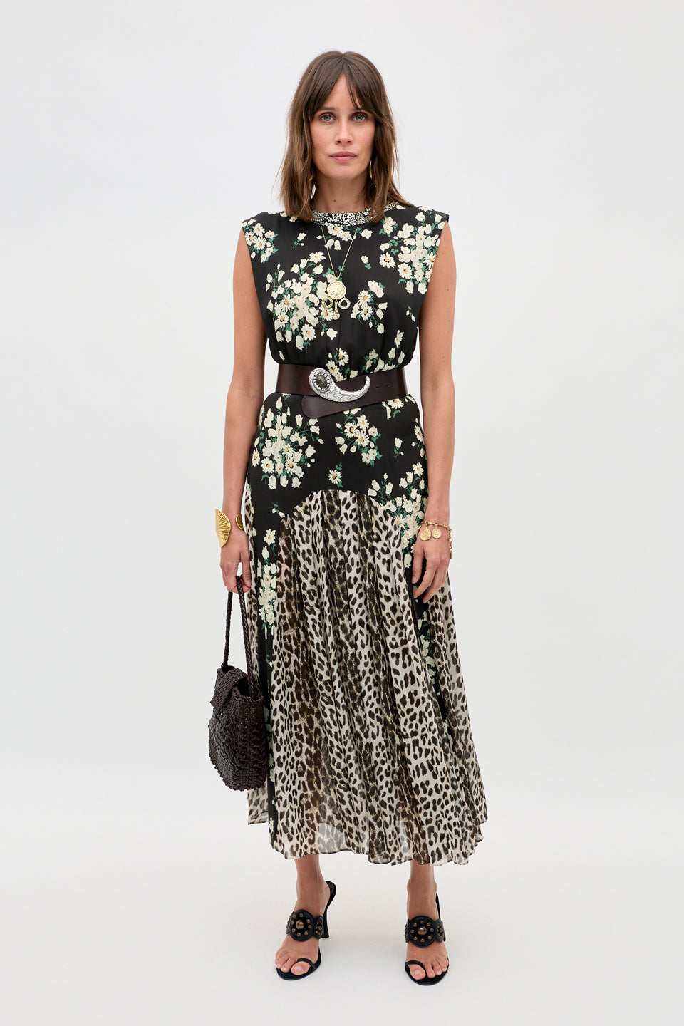 A woman stands against a plain background wearing the Coralie - Amelia Gremlin Mix Black top, paired with a pleated leopard print skirt, black sandals, jewelry, a belt, and holding a woven handbag.