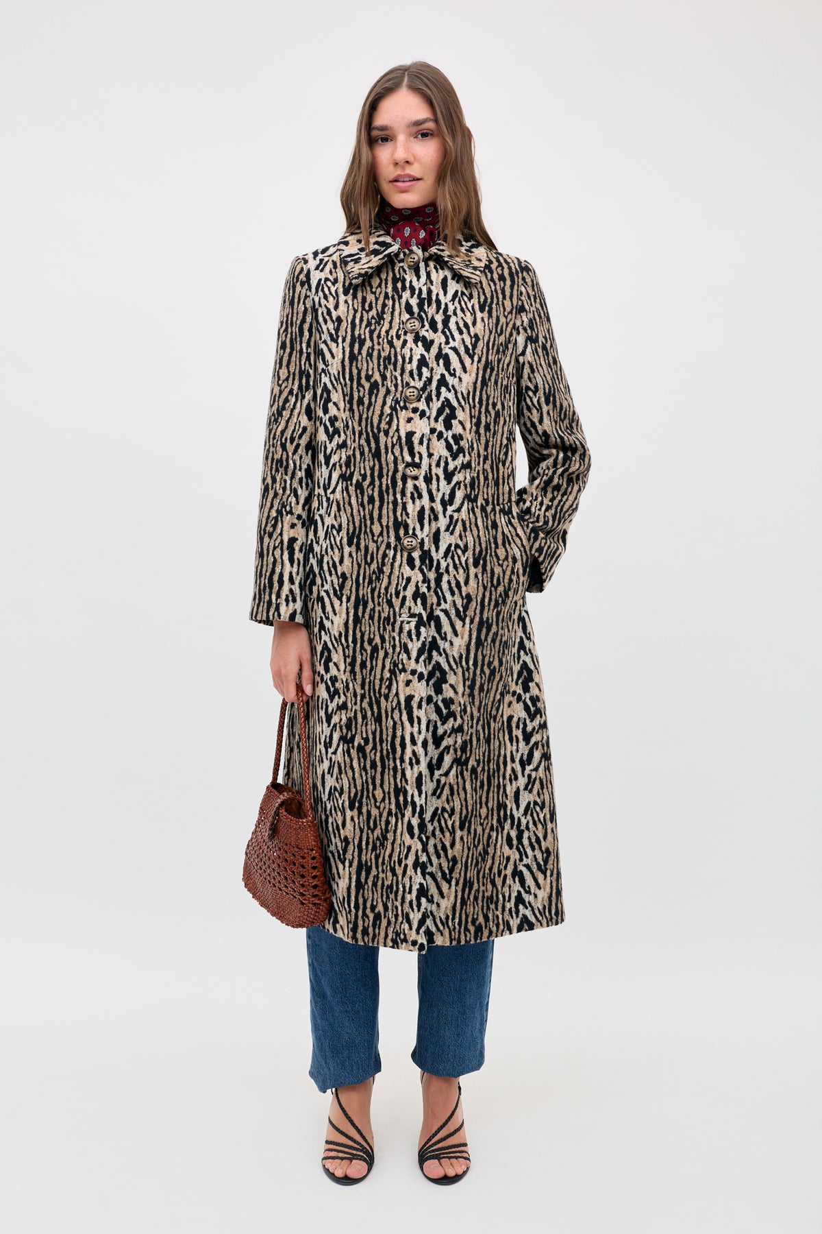A woman in the Milly - Bohemia Leopard coat over blue jeans and black strappy sandals holds a brown woven handbag, facing the camera against a plain white background.