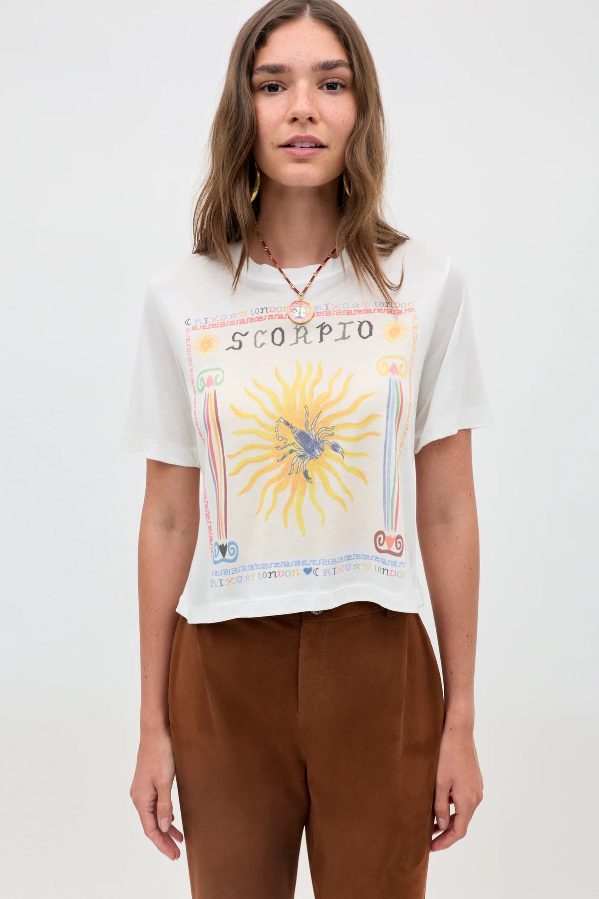 A woman with light brown hair wears the Ria - Scorpio white sheer jersey T-shirt, featuring a sun and scorpion illustration, paired with brown pants and a cropped hem, standing against a plain light background.