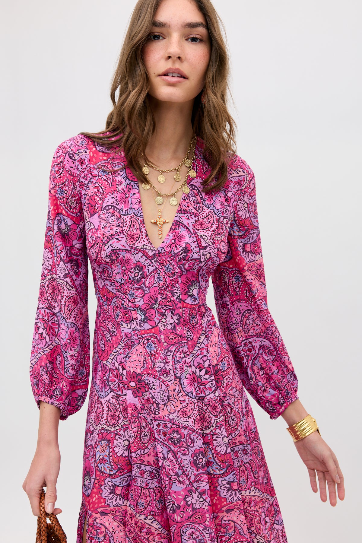 A woman wears the Camellia - Paisley Floral Red long-sleeved dress with a V-neck, layered gold necklaces, and a gold bracelet. She stands against a plain light background, holding a small brown bag.