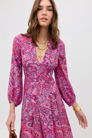 A woman wears the Camellia - Paisley Floral Red long-sleeved dress with a V-neck, layered gold necklaces, and a gold bracelet. She stands against a plain light background, holding a small brown bag.
