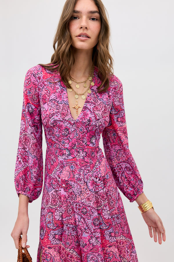 A woman wears the Camellia - Paisley Floral Red long-sleeved dress with a V-neck, layered gold necklaces, and a gold bracelet. She stands against a plain light background, holding a small brown bag.