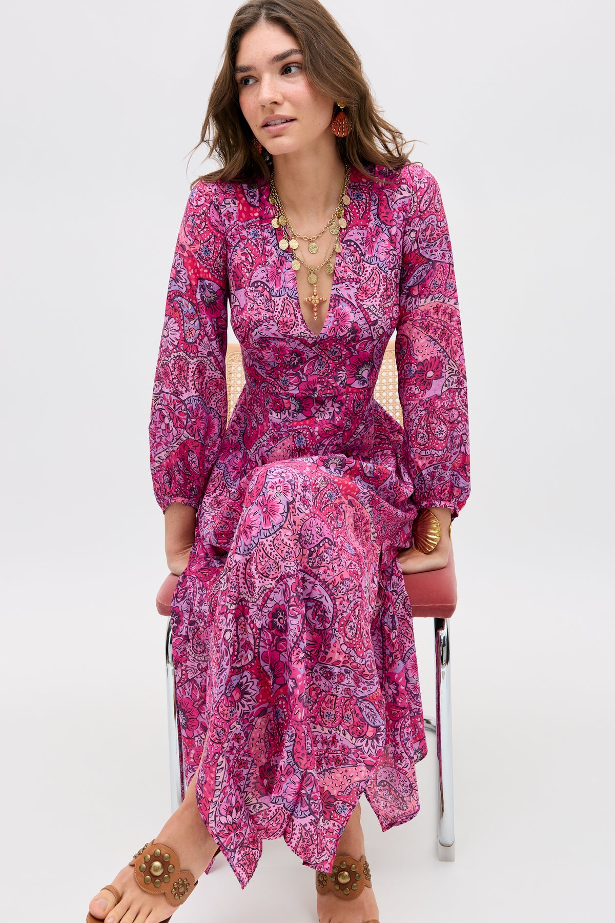 A woman sits on a pink chair, wearing the Camellia - Paisley Floral Red long-sleeved flowy dress, styled with layered necklaces, large earrings, and sandals, against a plain light background.