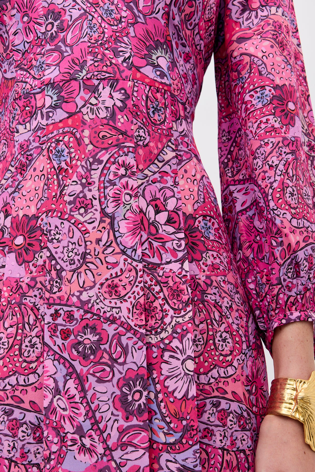 A close-up of a person wearing the Camellia - Paisley Floral Red long-sleeved dress, featuring vibrant pink, purple, and red florals and paisleys, paired with a chunky gold bracelet.