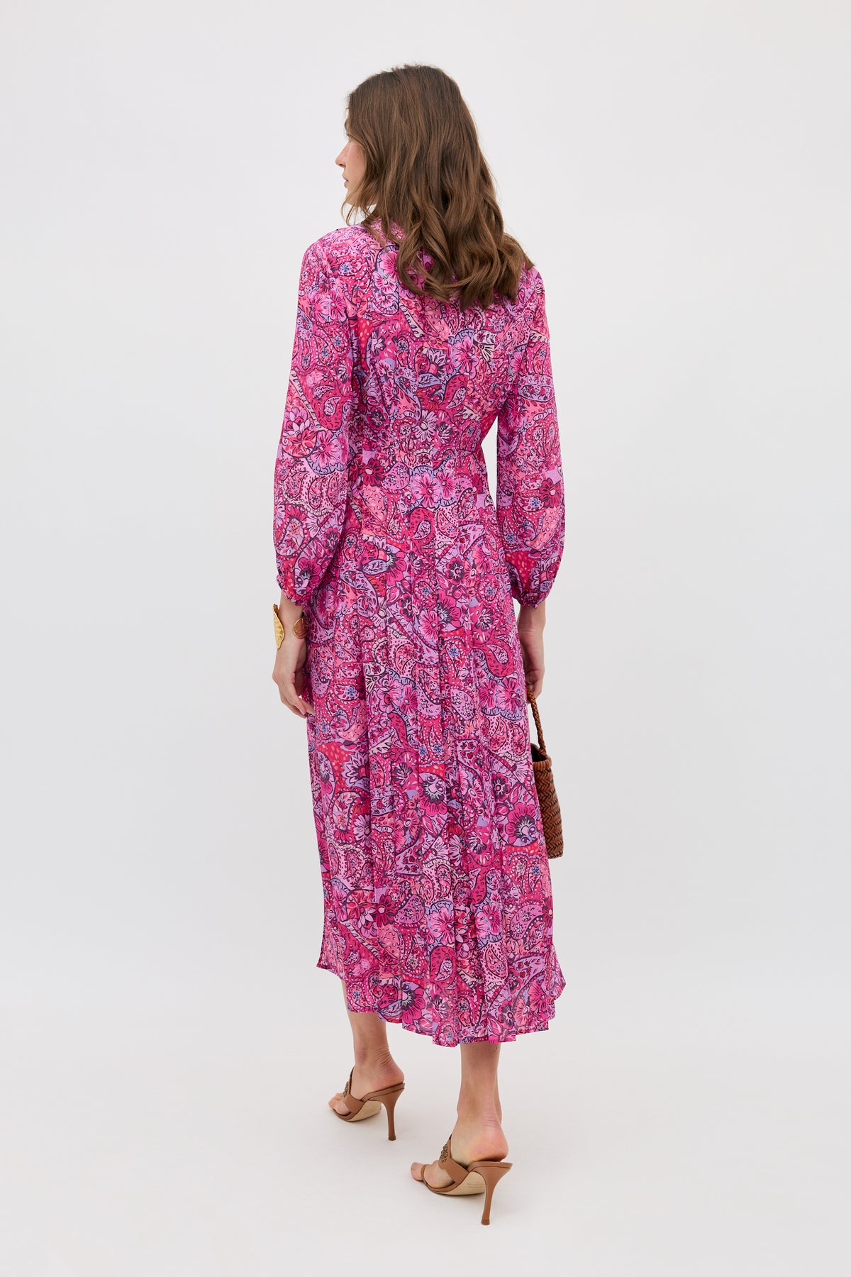 A woman faces away in the Camellia - Paisley Floral Red long-sleeved, ankle-length dress with a flowy skirt. She holds a brown handbag and wears tan heeled sandals against a plain white background.