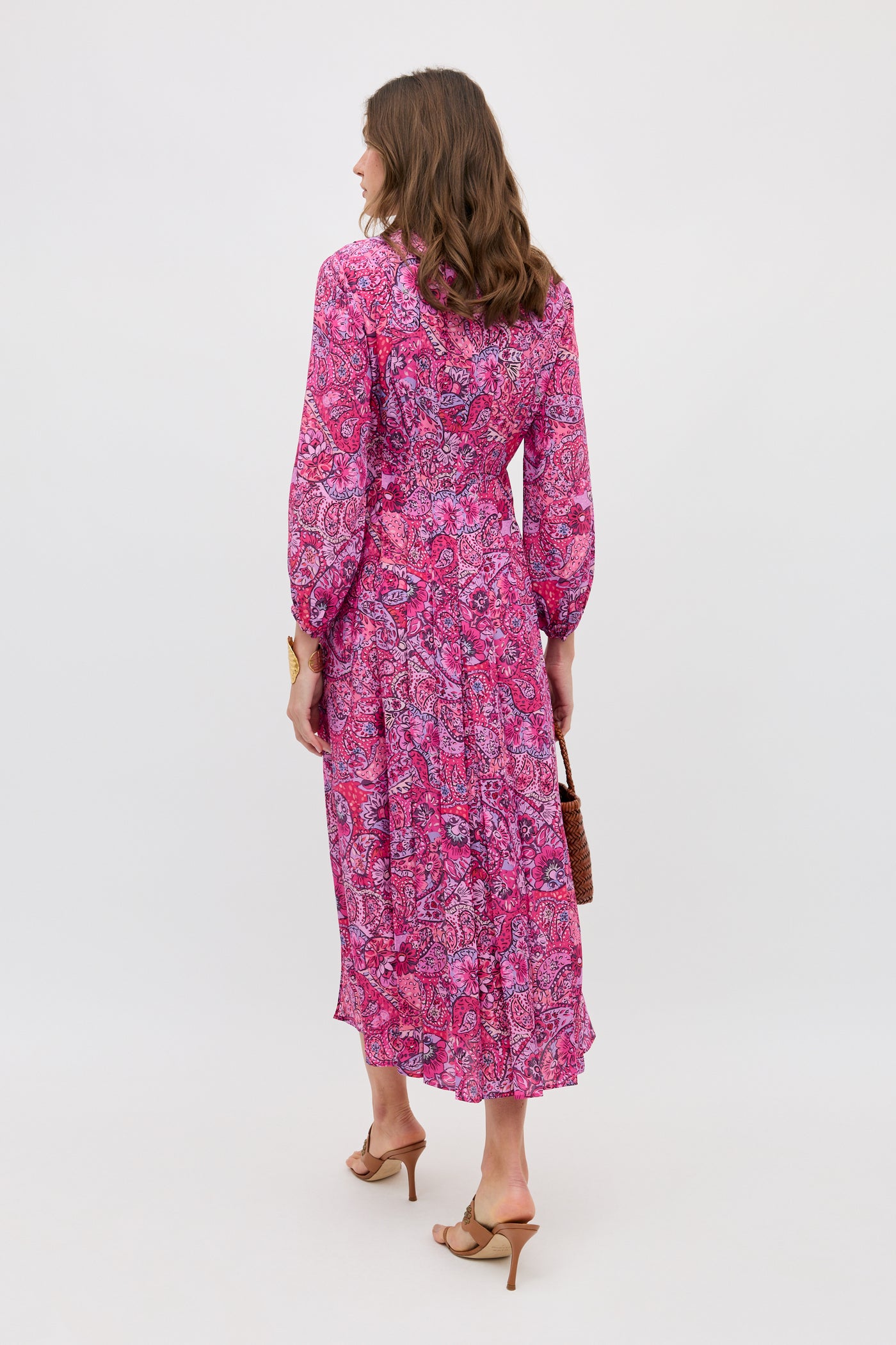 A woman faces away in the Camellia - Paisley Floral Red long-sleeved, ankle-length dress with a flowy skirt. She holds a brown handbag and wears tan heeled sandals against a plain white background.