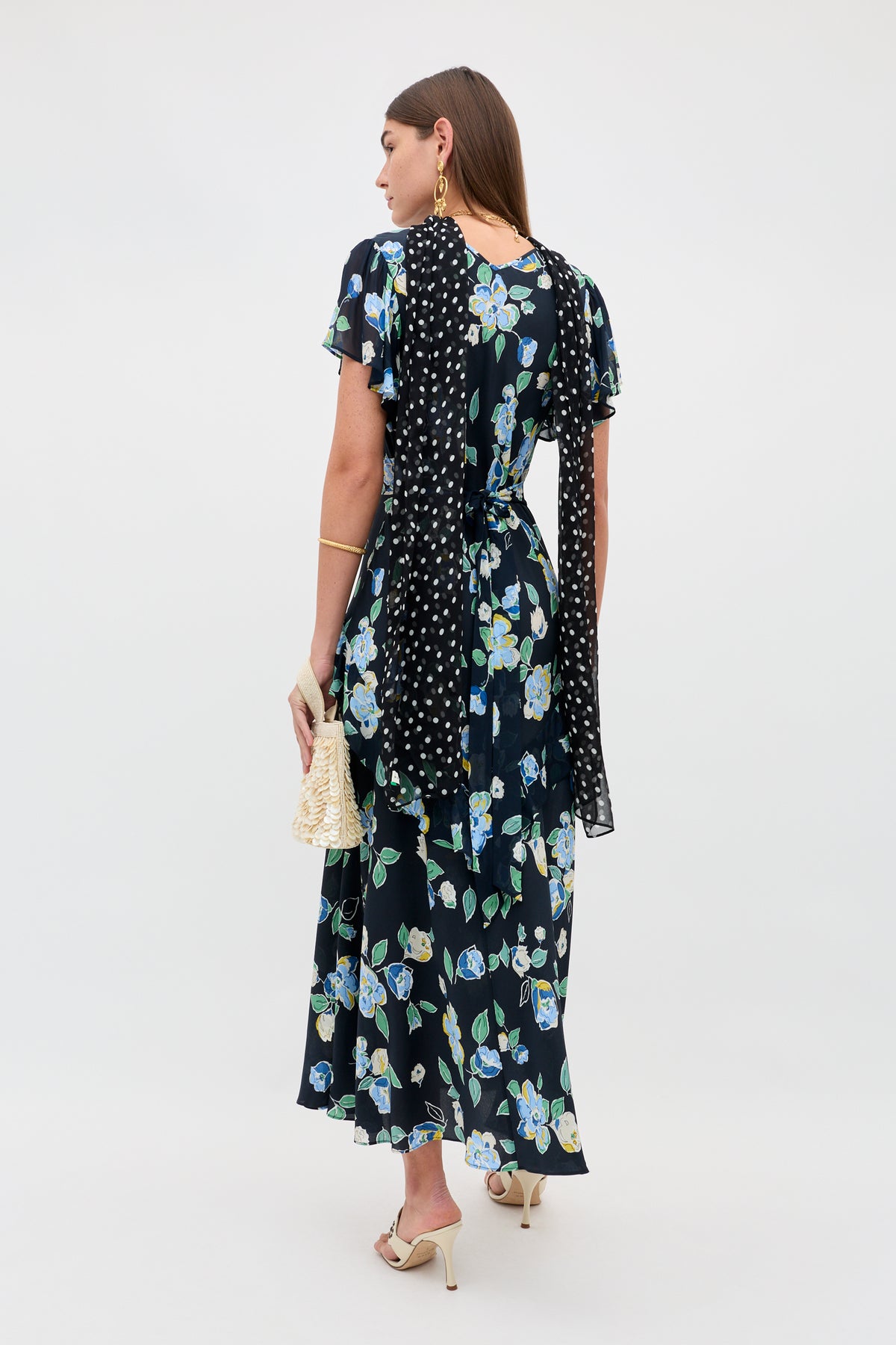 A woman with long brown hair wears the Evie - Kimmie Floral Navy dress, a black scarf with white polka dots, cream heeled sandals, gold jewelry, and carries a woven handbag.