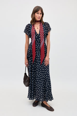 A woman stands against a plain background wearing the Evie - Bohemia Spot Deep Navy dress. She pairs it with a maroon printed scarf, a woven brown bag, and matching woven brown flats.