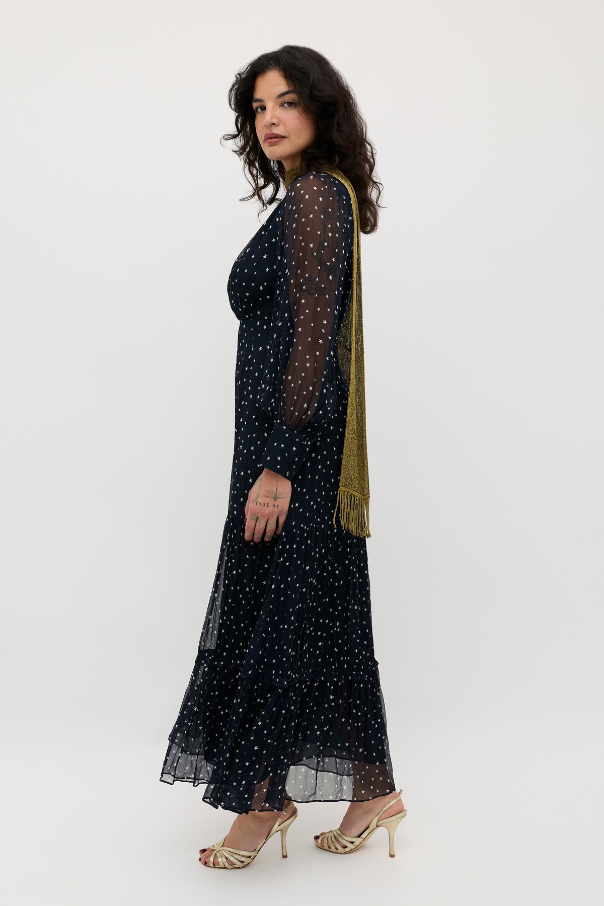 A woman with wavy dark hair, wearing the long, sheer navy Elsie - Starry Night dress with white polka dots, a gold tasseled scarf, and gold strappy heels stands in profile against a plain white background.