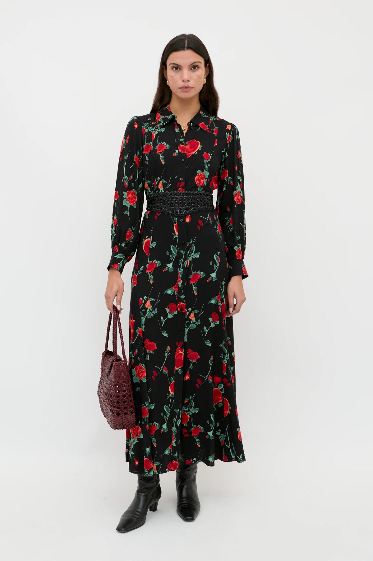 A woman with long brown hair and a neutral expression stands against a white background wearing the Maddison - Claudia Rose Black, styled with black boots and a dark red woven handbag for a chic everyday look.