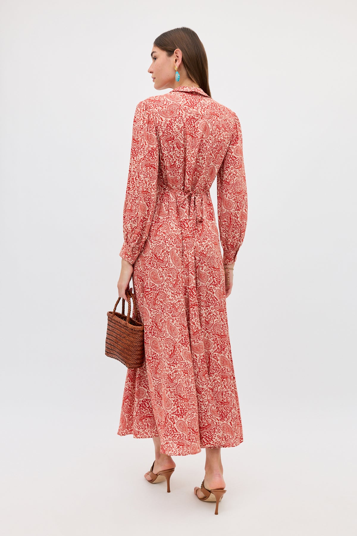 Woman standing and facing away, wearing the Maddison - Woodcut Gremlin Red long-sleeved dress with a cinched waist, tan heeled sandals, turquoise earrings, and holding a brown woven handbag.