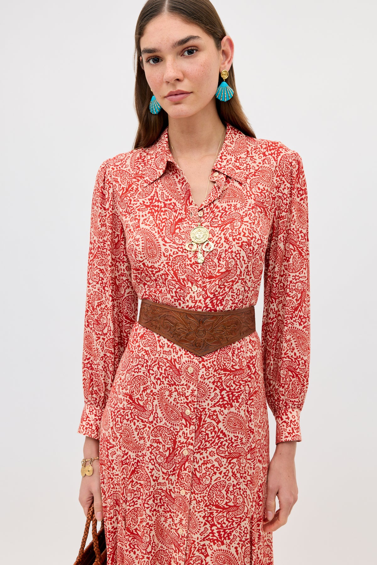 A woman stands against a plain background wearing the Maddison - Woodcut Gremlin Red dress, styled with a brown wide belt, turquoise tasseled earrings, layered gold necklaces, and holding a brown woven bag.