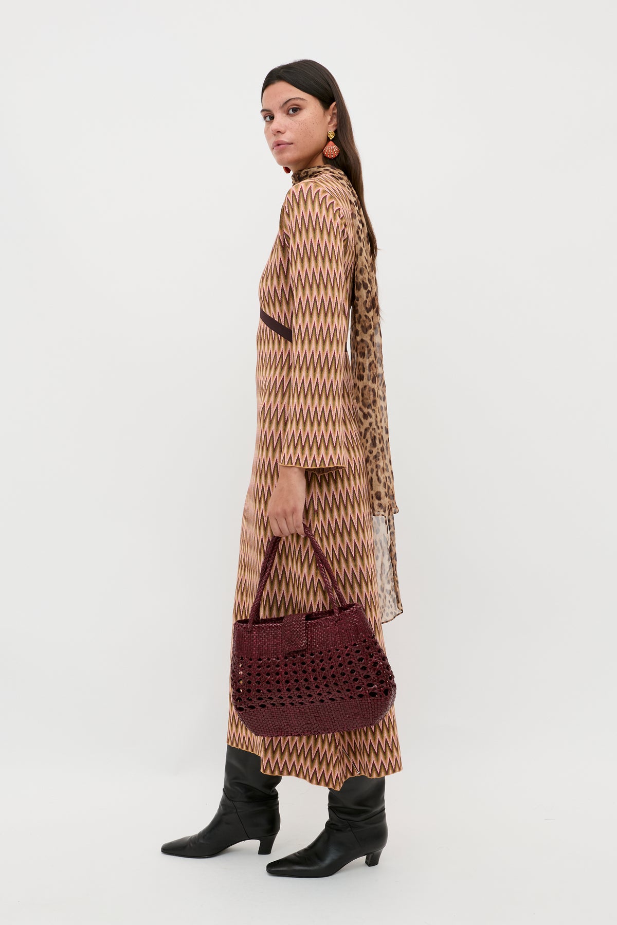 A woman models the Pamela - Maeve Jacquard Multi dress, featuring zigzag patterns. She stands sideways with a dark red woven bag, black boots, statement earrings, and a draped scarf against a white background.