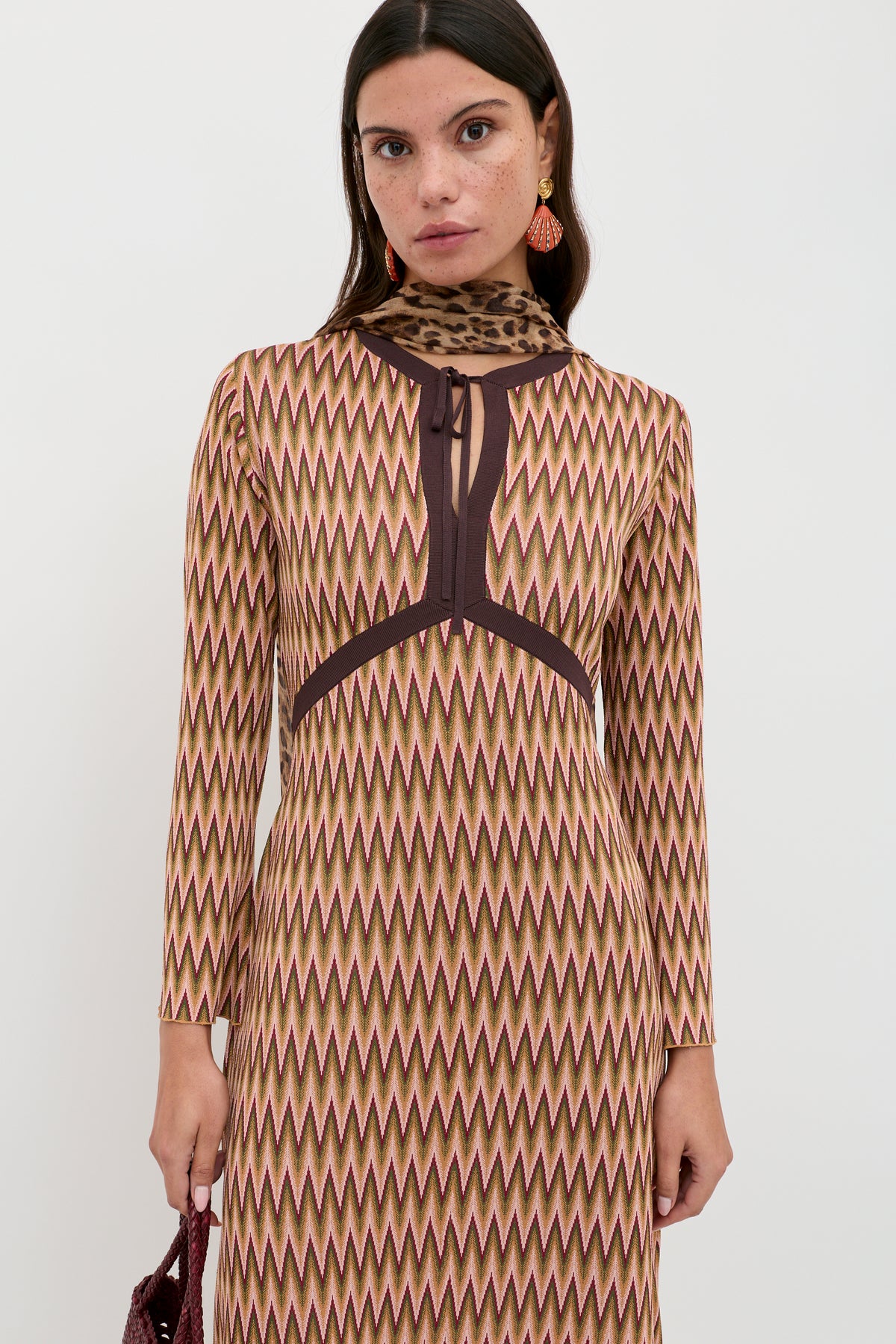 A woman wearing the Pamela - Maeve Jacquard Multi dress, featuring long sleeves and an earthy zigzag pattern, stands against a white background. She has straight brown hair and accessorizes with a brown animal-print scarf.