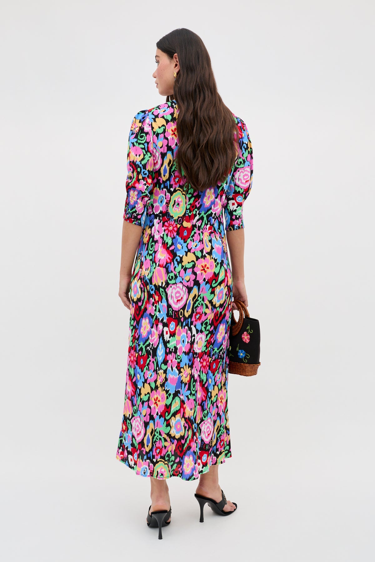 A woman with long brown hair faces away, wearing the Jess - Ikat Bloom Charcoal dress with black heeled sandals and a small black handbag. The background is plain and light.