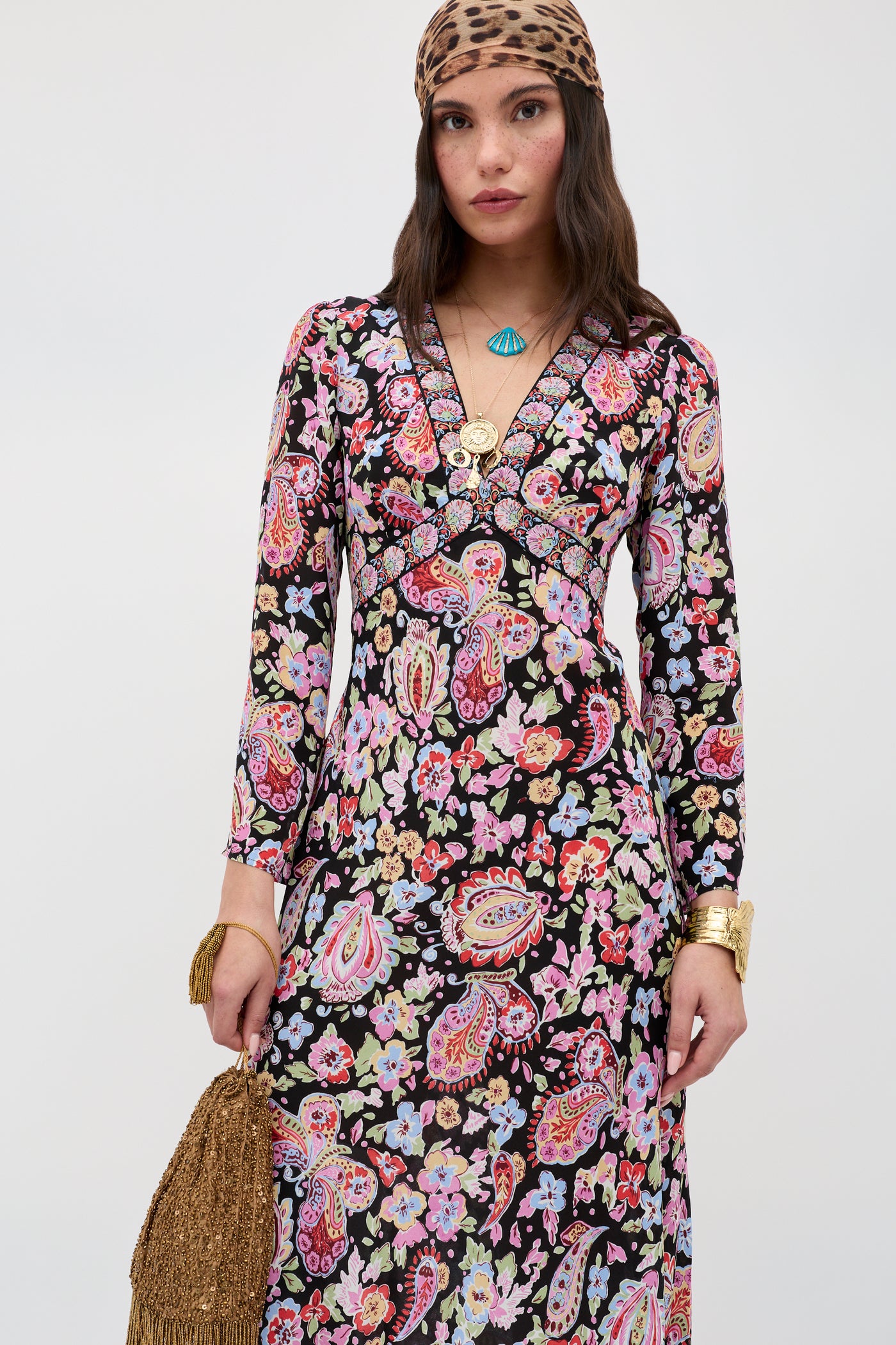 A woman wears the Tania - Cara Floral Black long-sleeve V-neck dress with a colorful paisley and floral pattern, paired with a leopard print headscarf, gold jewelry, and a fringed bag against a plain light background.