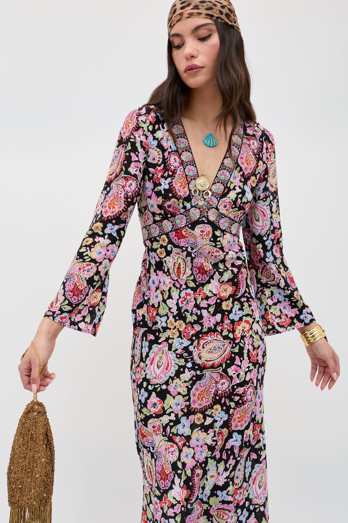 A woman poses against a plain background wearing the Tania - Cara Floral Black dress, a long-sleeved V-neck with vibrant floral and paisley patterns, accessorized with a leopard-print headband, layered necklaces, gold bracelets, and a fringed gold bag.