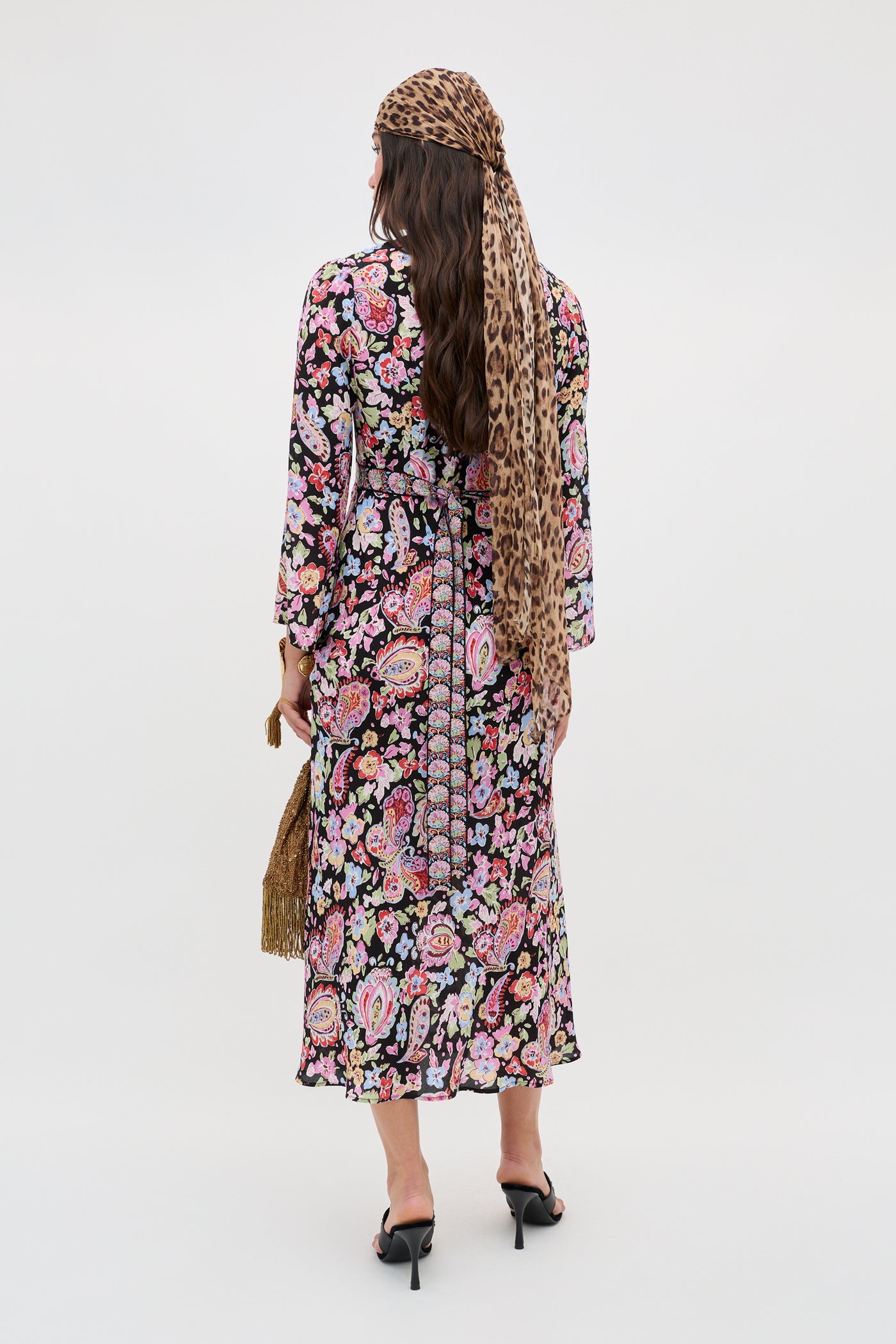 A woman faces away, wearing the Tania - Cara Floral Black long-sleeve dress, paired with black heels, a cheetah print headscarf, and a tan fringed bag against a plain light background.