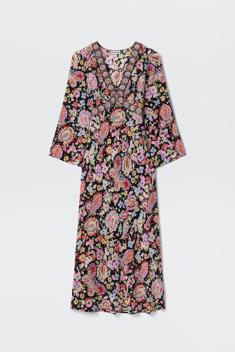 The Tania - Cara Floral Black is a long-sleeved V-neck maxi dress with a vibrant floral and paisley pattern on a black background, shown flat on a white surface.
