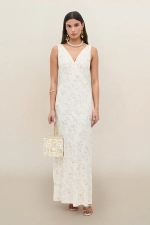 A woman wears the Fawn - Botanical Embroidery Pearl sleeveless, floor-length white dress with subtle floral patterns, styled with a woven handbag, gold jewelry, and heeled sandals against a light background.