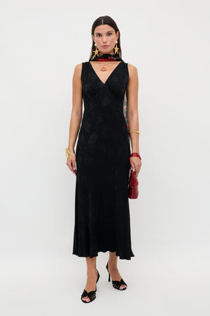 A woman poses against a white background in the Sandrine - Black Poppy Jacquard slip dress, featuring a sleeveless, V-neck, bias-cut silhouette with a subtle poppy pattern. She pairs it with bold jewelry, a red handbag, and black open-toe heels.
