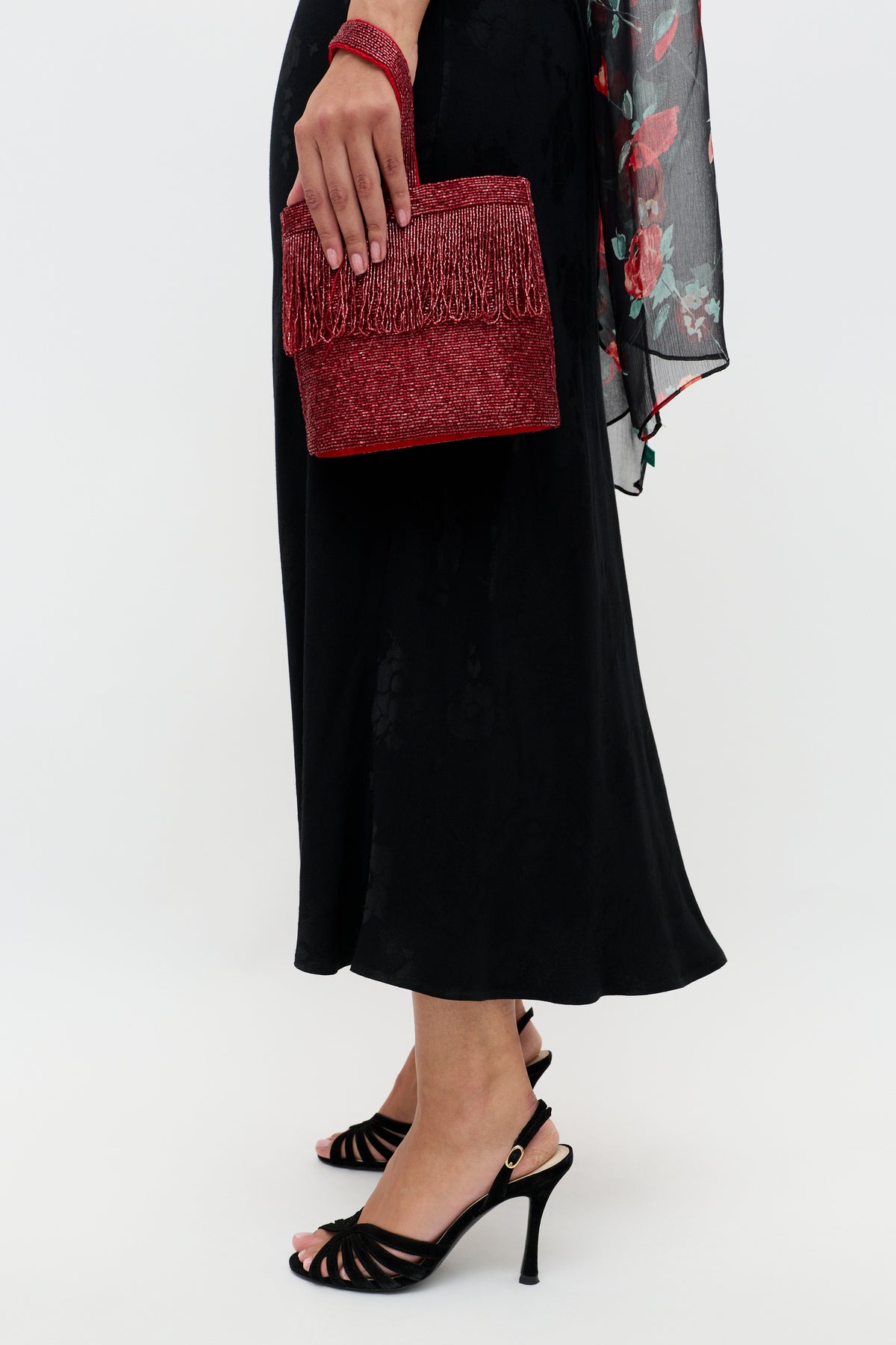 A person in the Sandrine - Black Poppy Jacquard dress and black heeled sandals holds a red, glittery fringe handbag against a plain backdrop.