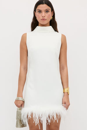 A woman wears the Candice dress—sleeveless, sixties-inspired, in satin back silk crepe with an ostrich-feather hem—posing against a plain background. She carries a small gold bag and accessorizes with gold jewelry.