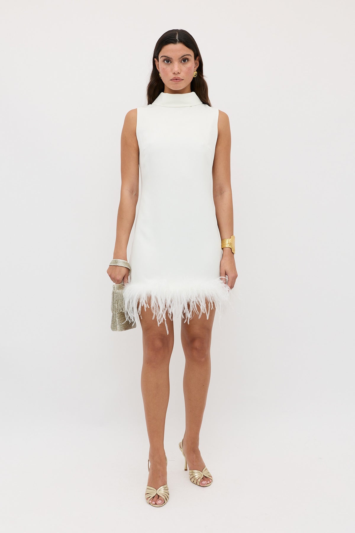 A woman stands against a plain white background wearing the Candice - Satin Back Silk Crepe, a sixties-inspired shift dress with a high neckline and ostrich-feathered hemline, styled with gold heels, a gold bracelet, and a metallic clutch.