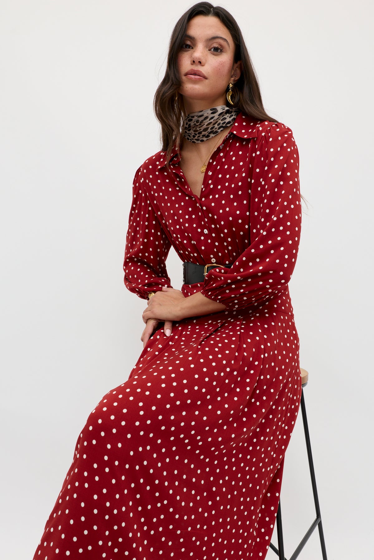 A woman sits on a stool wearing the Izzy - Layla Dot Red dress, featuring long sleeves and white polka dots, styled with a black belt and leopard print scarf, against a plain white background.