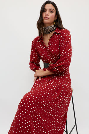 A woman sits on a stool wearing the Izzy - Layla Dot Red dress, featuring long sleeves and white polka dots, styled with a black belt and leopard print scarf, against a plain white background.