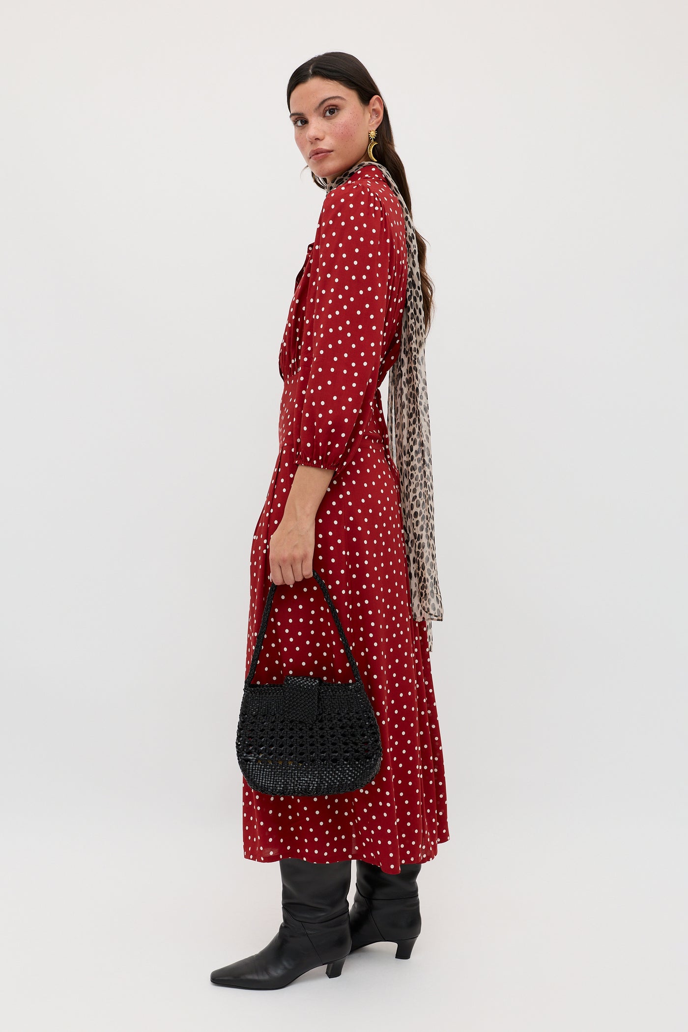 A woman stands sideways in the Izzy - Layla Dot Red long polka dot dress, black boots, a light animal-print scarf, and holding a black woven handbag. She has long dark hair against a plain white background.
