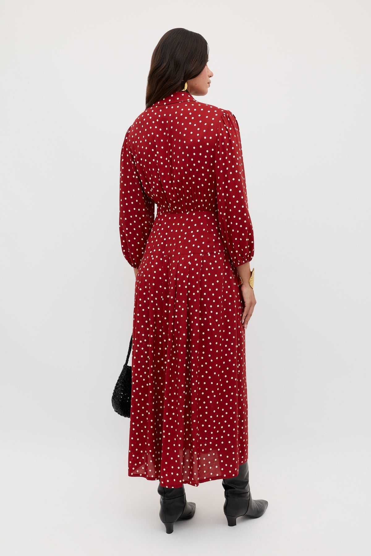 A woman with dark hair, facing away against a plain light background, wears the Izzy - Layla Dot Red long-sleeved ankle-length red dress with white polka dots, black boots, and carries a black textured handbag.