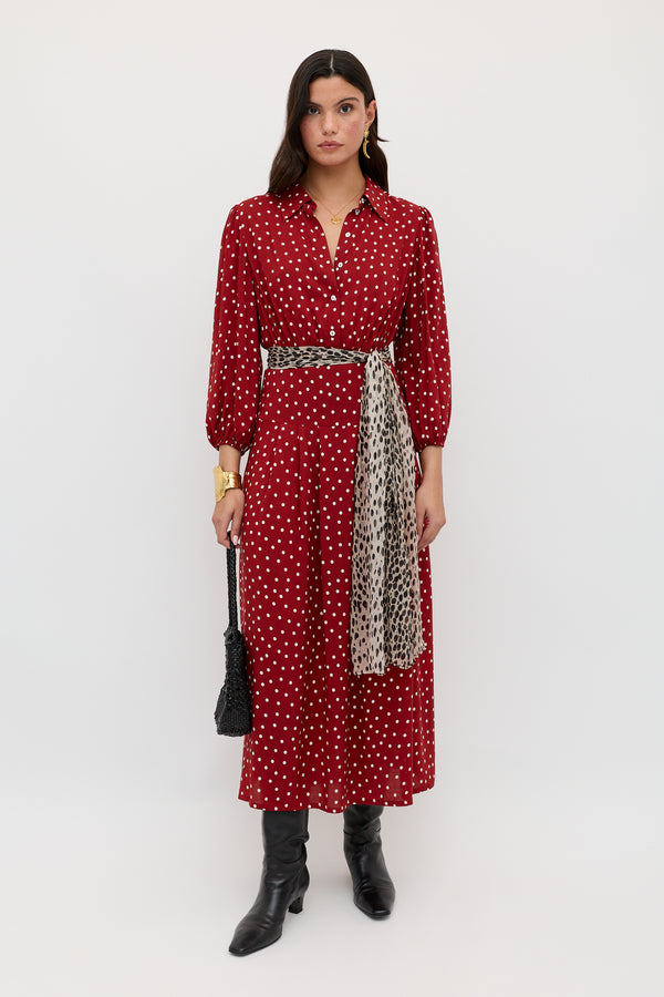 A woman stands against a white background wearing the Izzy - Layla Dot Red dress, paired with black boots, a black textured bag, a gold bracelet, and a patterned scarf tied at her waist.