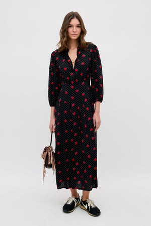 A woman with wavy hair and a relaxed expression stands against a plain background wearing the Izzy - Rose Spot Black long dress with red polka dots, black sneakers, and holding a small striped drawstring bag.