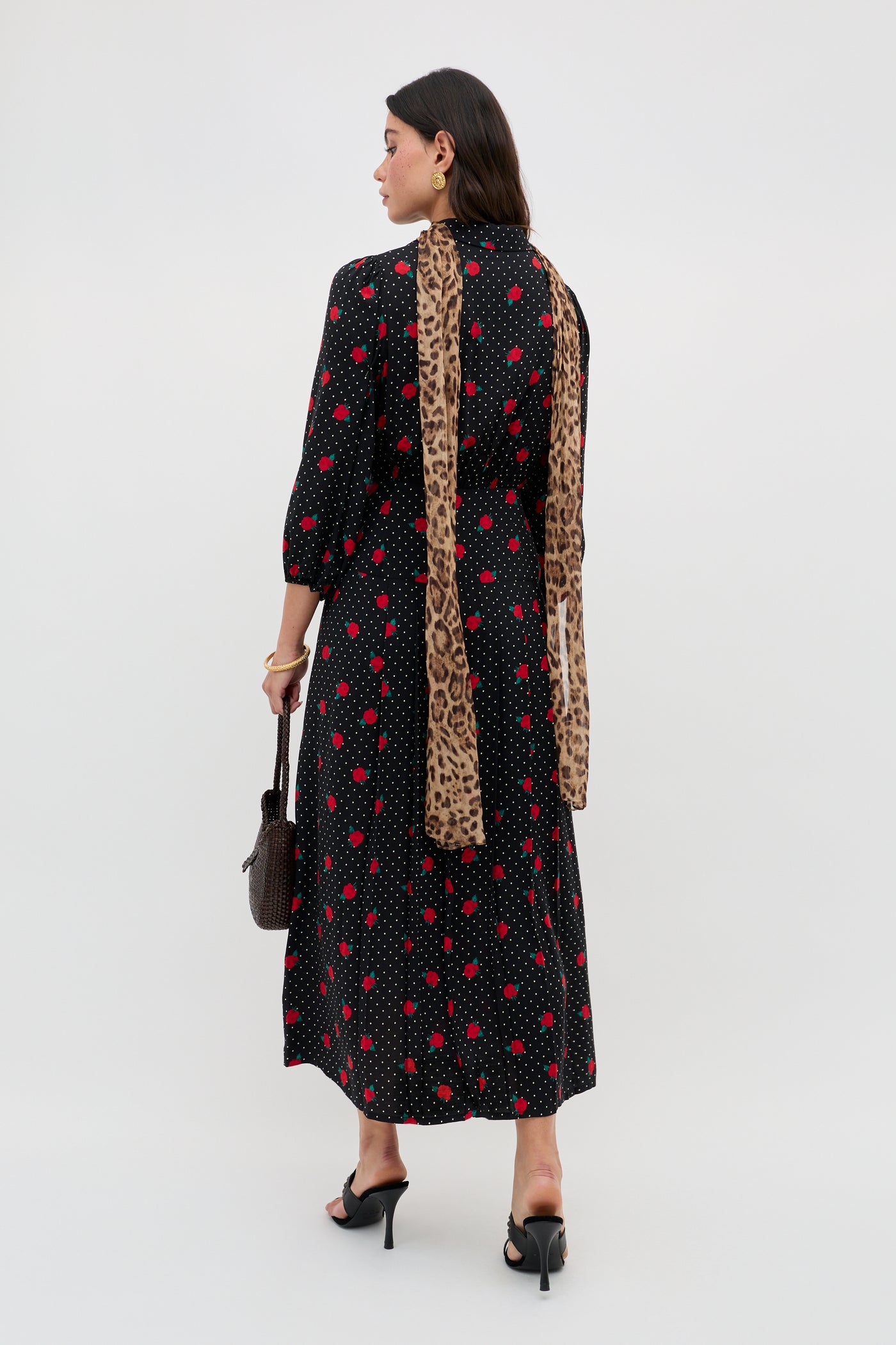 A woman wearing the Izzy - Rose Spot Black long-sleeved, ankle-length dress stands facing away, styled with black heels, a long leopard-print scarf, and a brown handbag.