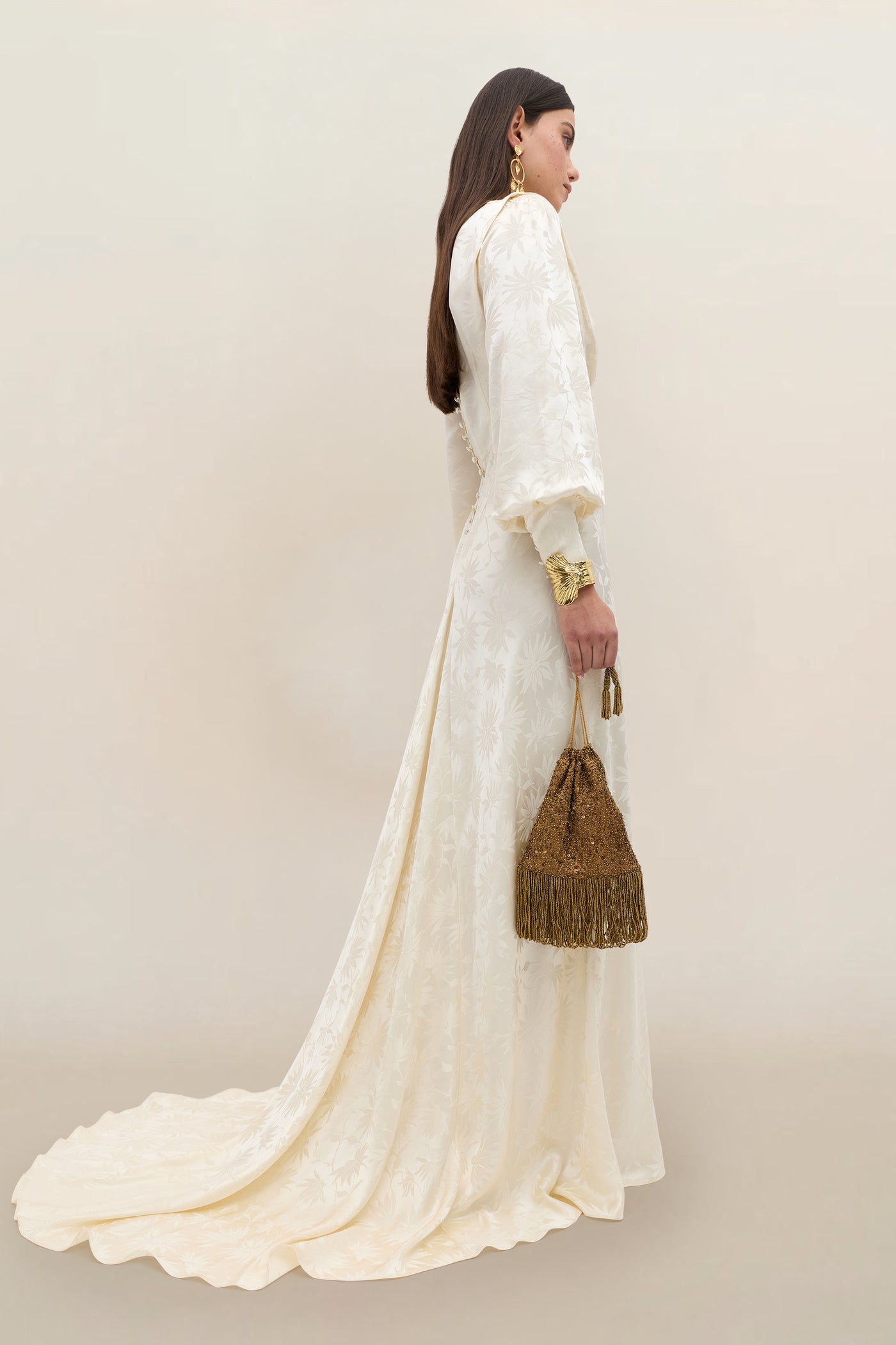 A woman stands sideways against a plain, light background, wearing the Clementine - Daisy Jacquard Champagne gown—a long, white floral-patterned silk jacquard dress with puffed sleeves. She holds a gold tasseled handbag and accessorizes with large earrings, bracelets, and her long brown hair flowing loosely.