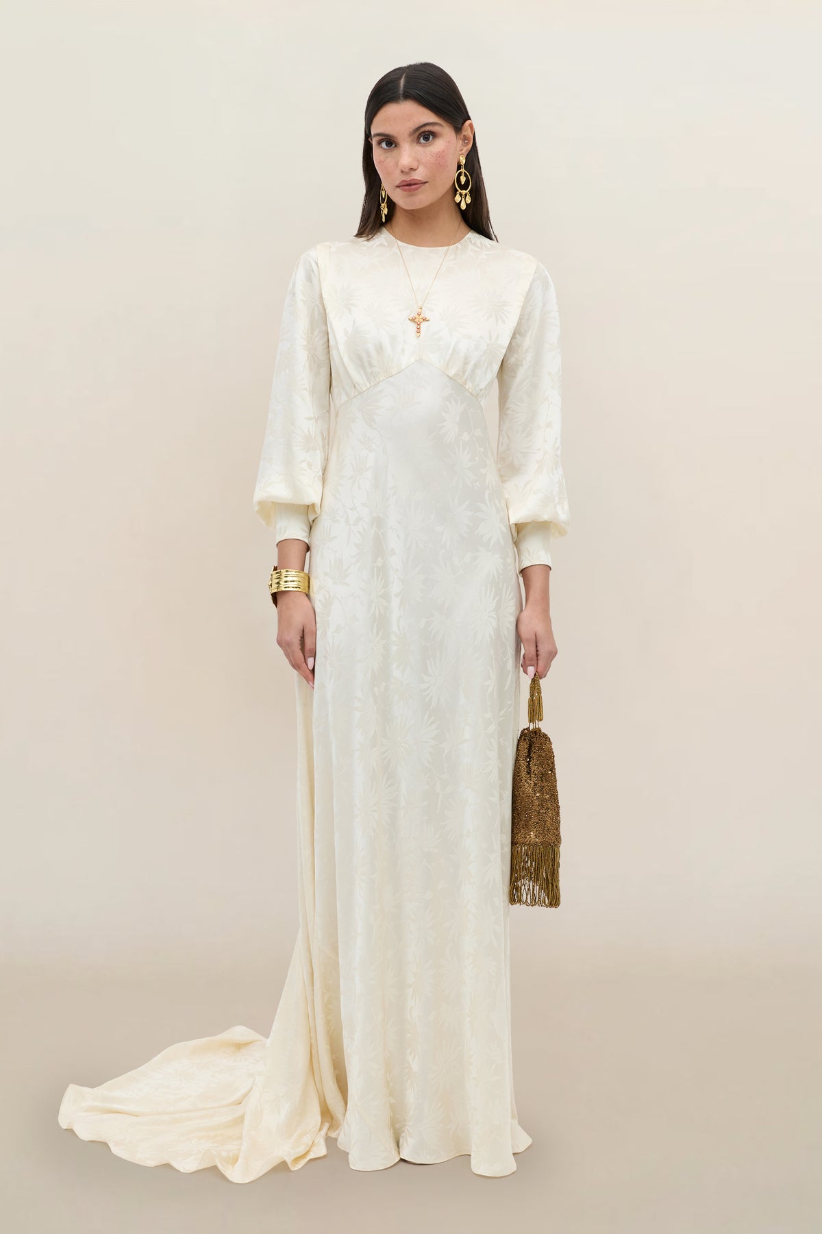 A woman in the Clementine - Daisy Jacquard Champagne gown, featuring a long ivory silk jacquard design with puffed sleeves, stands against a neutral background. She accessorizes with a gold fringed handbag and gold jewelry, including earrings, bracelets, and layered necklaces.