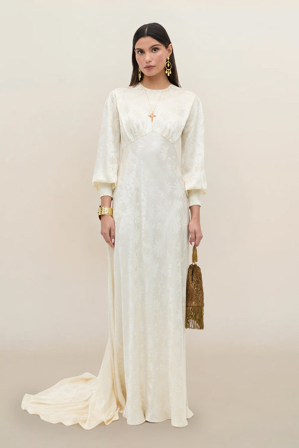 A woman in the Clementine - Daisy Jacquard Champagne gown, featuring a long ivory silk jacquard design with puffed sleeves, stands against a neutral background. She accessorizes with a gold fringed handbag and gold jewelry, including earrings, bracelets, and layered necklaces.
