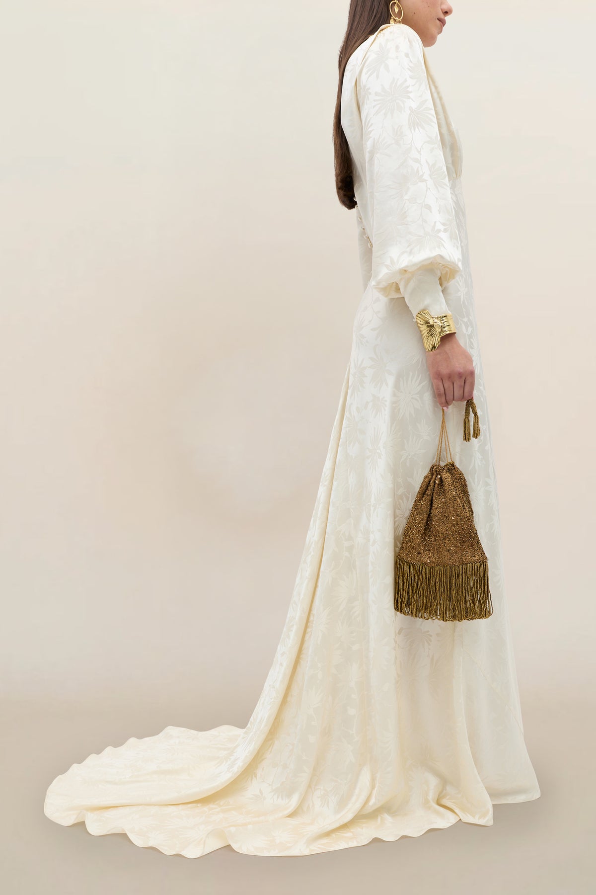 A woman wears the Clementine - Daisy Jacquard Champagne gown, a long, champagne-colored silk jacquard dress with a train. She holds a gold fringe purse and accessorizes with gold earrings and a chunky bracelet. The soft beige background highlights the effortlessly elegant bridal gown look.