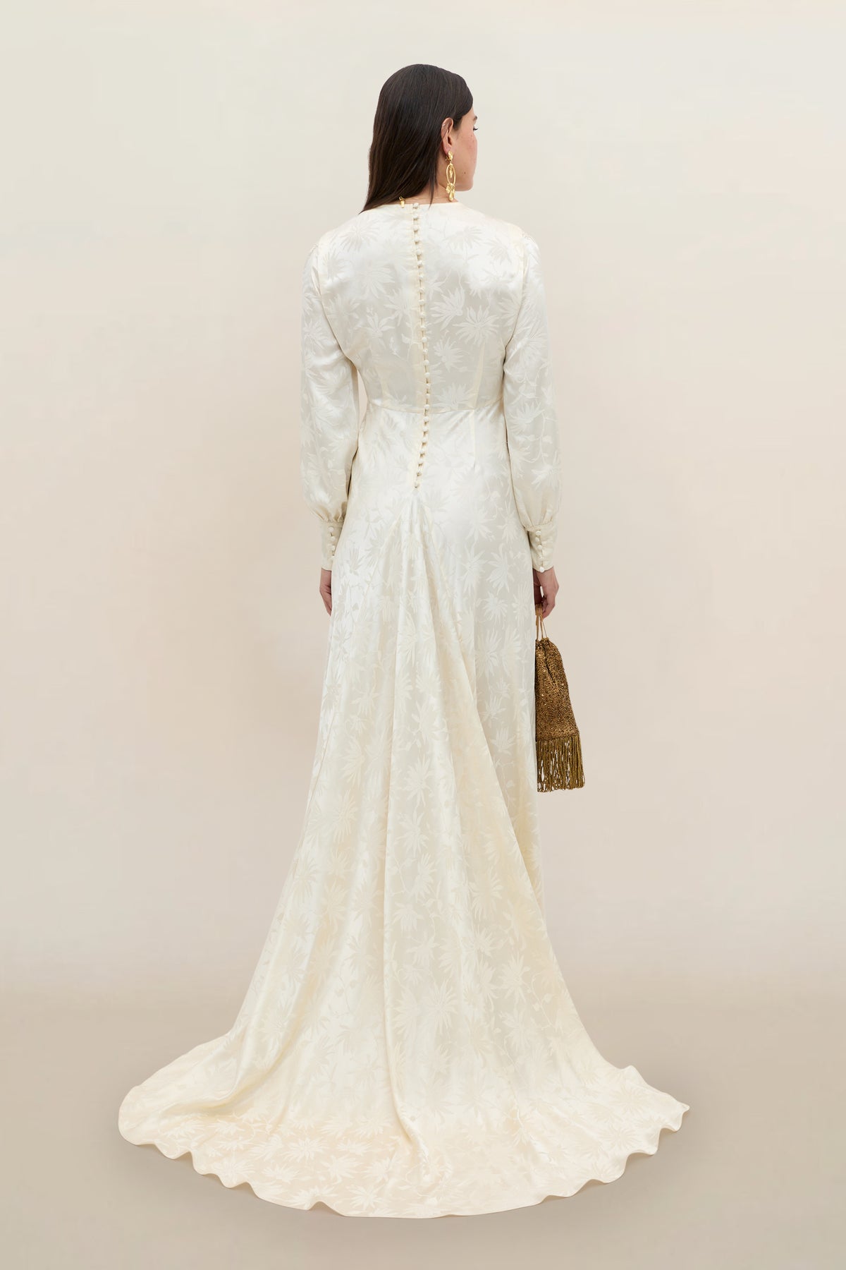 A woman in the Clementine - Daisy Jacquard Champagne gown, featuring long sleeves and an elegant floral jacquard pattern in ivory, stands facing away to reveal a row of back buttons and a subtle train. She accessorizes with a textured gold handbag and gold earrings.