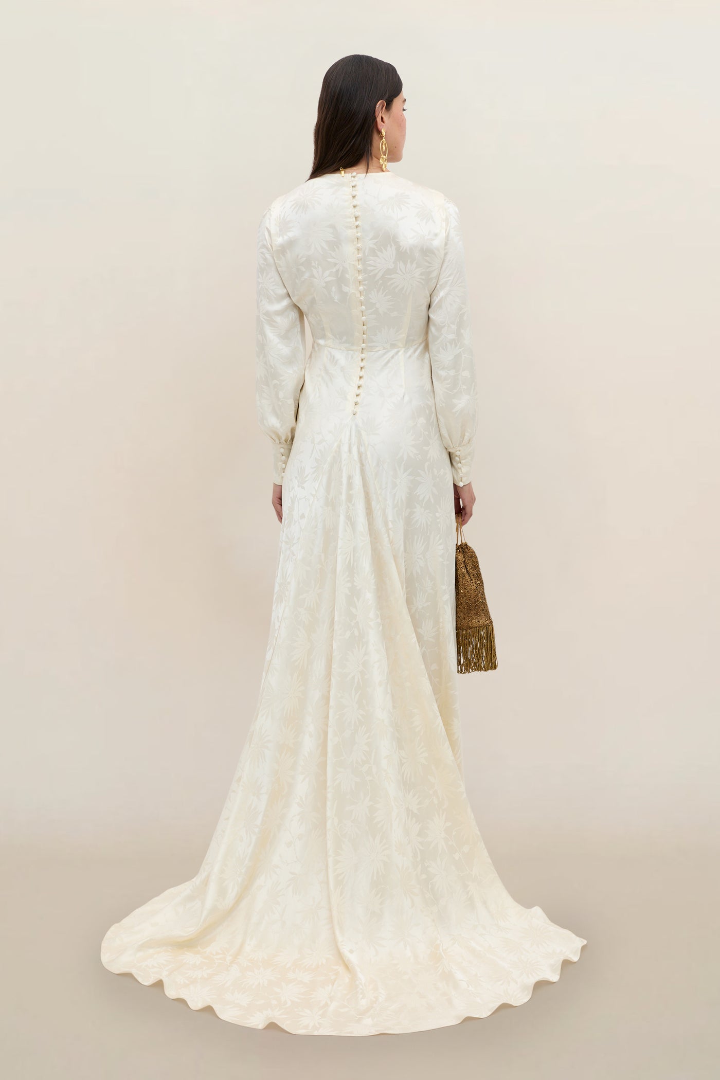 A woman in the Clementine - Daisy Jacquard Champagne gown, featuring long sleeves and an elegant floral jacquard pattern in ivory, stands facing away to reveal a row of back buttons and a subtle train. She accessorizes with a textured gold handbag and gold earrings.