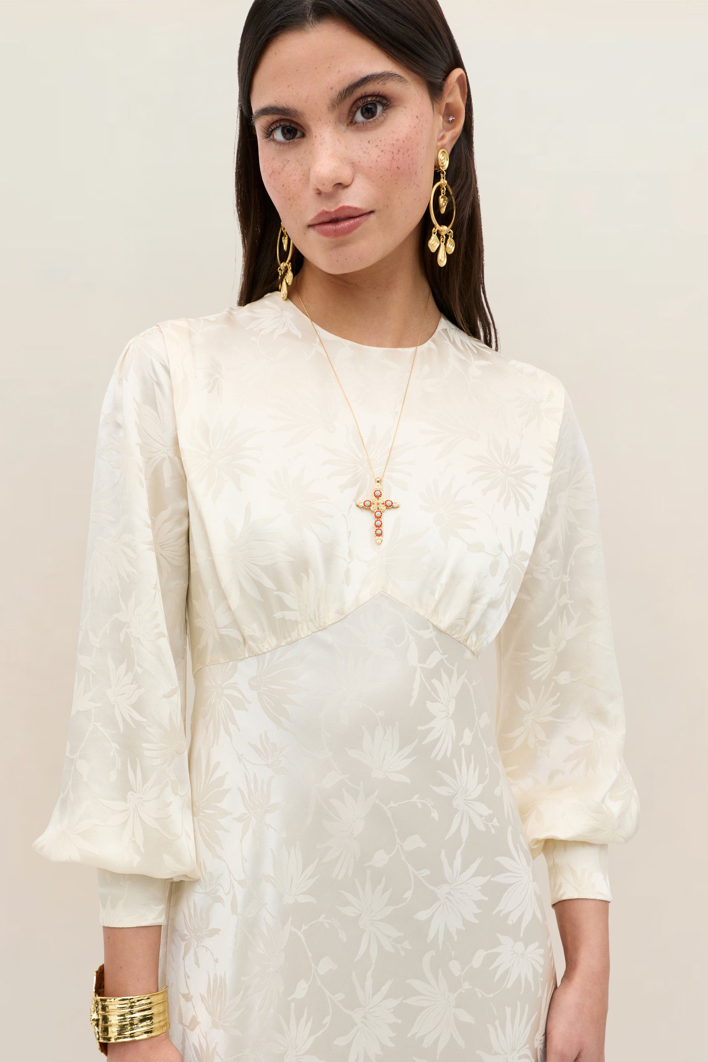 A woman stands against a plain beige background wearing the Clementine - Daisy Jacquard Champagne dress, an elegant cream floral-patterned piece reminiscent of a bridal gown. She accessorizes with gold earrings, a cross necklace, and a gold bracelet. With long dark hair, freckles, and a calm expression, she embodies understated sophistication.