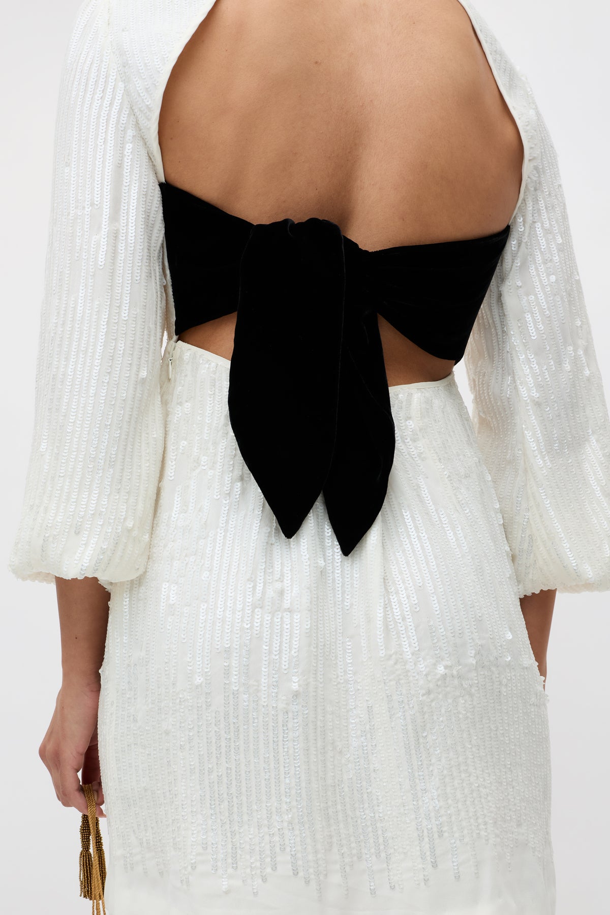 A person wearing the "lara - ivory black bow" dress, a white textured sequin design with an open back and a large black bow tied at the waist.