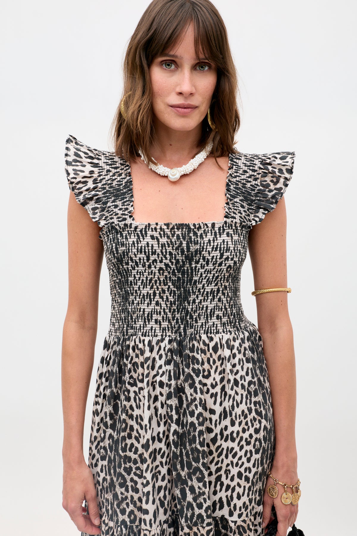 A woman with shoulder-length brown hair wears the Kendall - Nina Leopard Mocha dress with a ruffled, square neckline, accessorized with gold jewelry and a white shell necklace, standing against a plain backdrop.