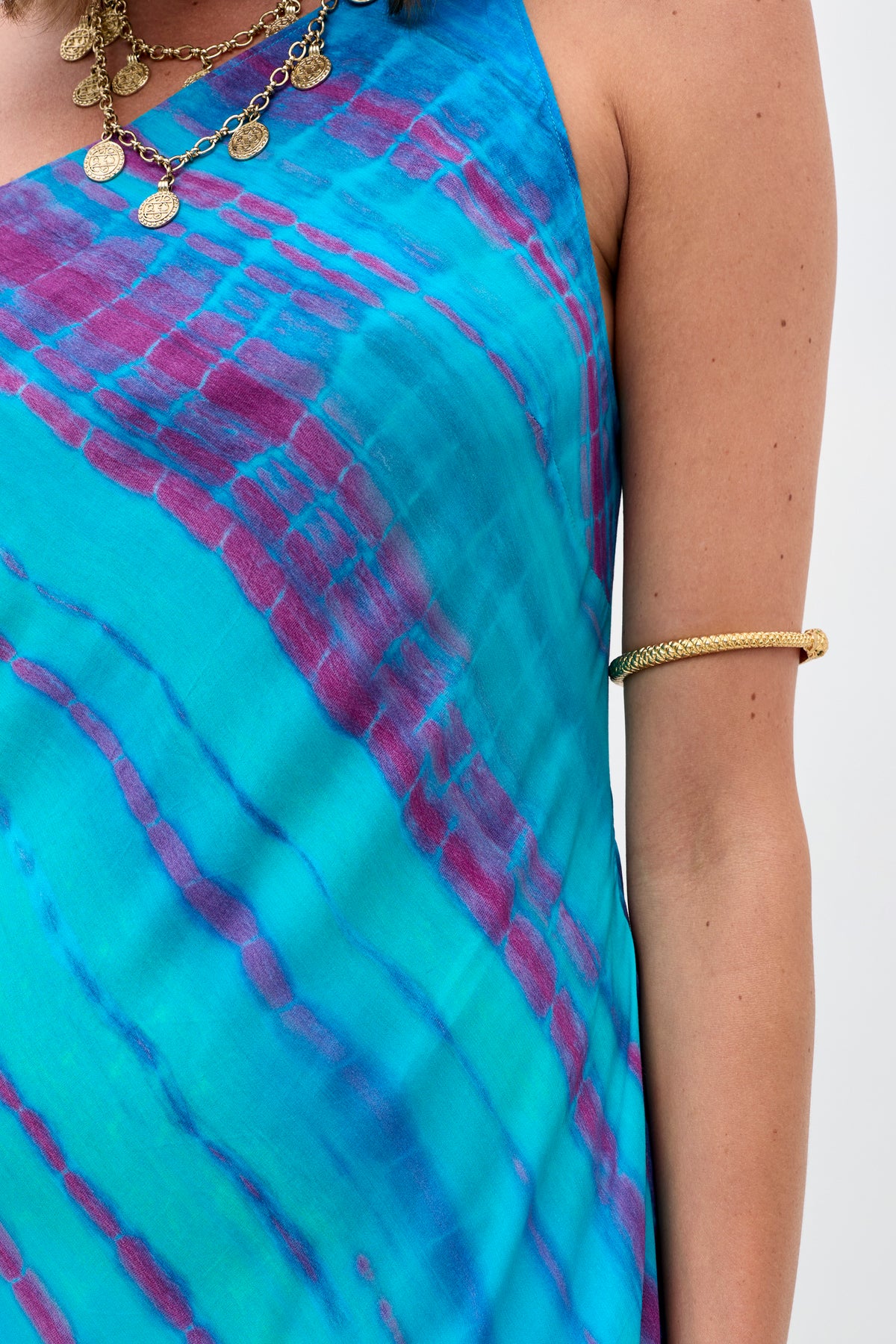 A person wearing the Doutzen - Tie Dye Blue dress, paired with a coin necklace and a gold arm cuff. Only the upper torso, shoulder, and part of the arm are visible against a plain background.