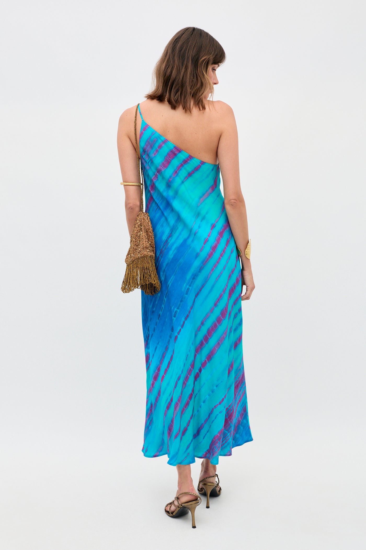 A woman stands facing away against a plain white background, wearing the Doutzen - Tie Dye Blue one-shoulder dress, styled with gold heels, gold accessories, and holding a gold fringed bag.