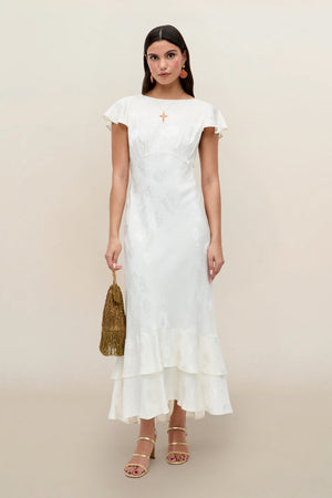 A woman wears the Liberty - Poppy ivory jacquard ankle-length dress with ruffle hem and cap sleeves, styled with gold earrings, strappy heels, and a gold fringed bag against a plain beige background.